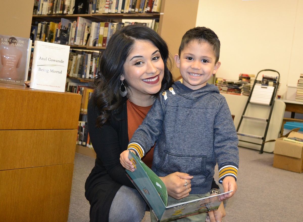 A woman smiles as she puts her arms around her son who is reading a book. She is wearing a sweater, top and jeans. He is wearing a sweatshirt. Behind them are bookshelves.