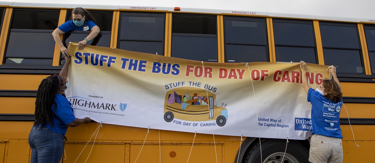 Three women hang a banner that says “Stuff the Bus for Day of Caring” from the windows of a school bus.
