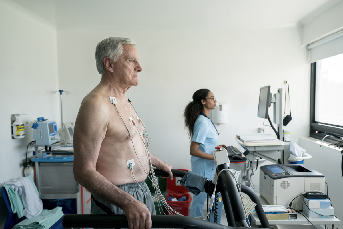 A man wears several electrodes on his chest as he walks on a treadmill. A woman in scrubs is in the background, working on a computer.