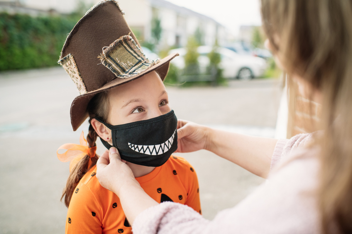 Young girl dressed for Halloween in brown top hat and pumpkin shirt gets her black smiley-face cloth facemask fitted by her mother prior to trick-or-treating.