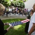 Two women fold the American flag. A crowd of people watch from a distance.