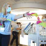 Dr. Sarah Iriana extends her arm holding a handful of ribbon next to a crib. Equipment is draped from the side of the crib. Behind her, three women in surgical masks look on or focus on other work.