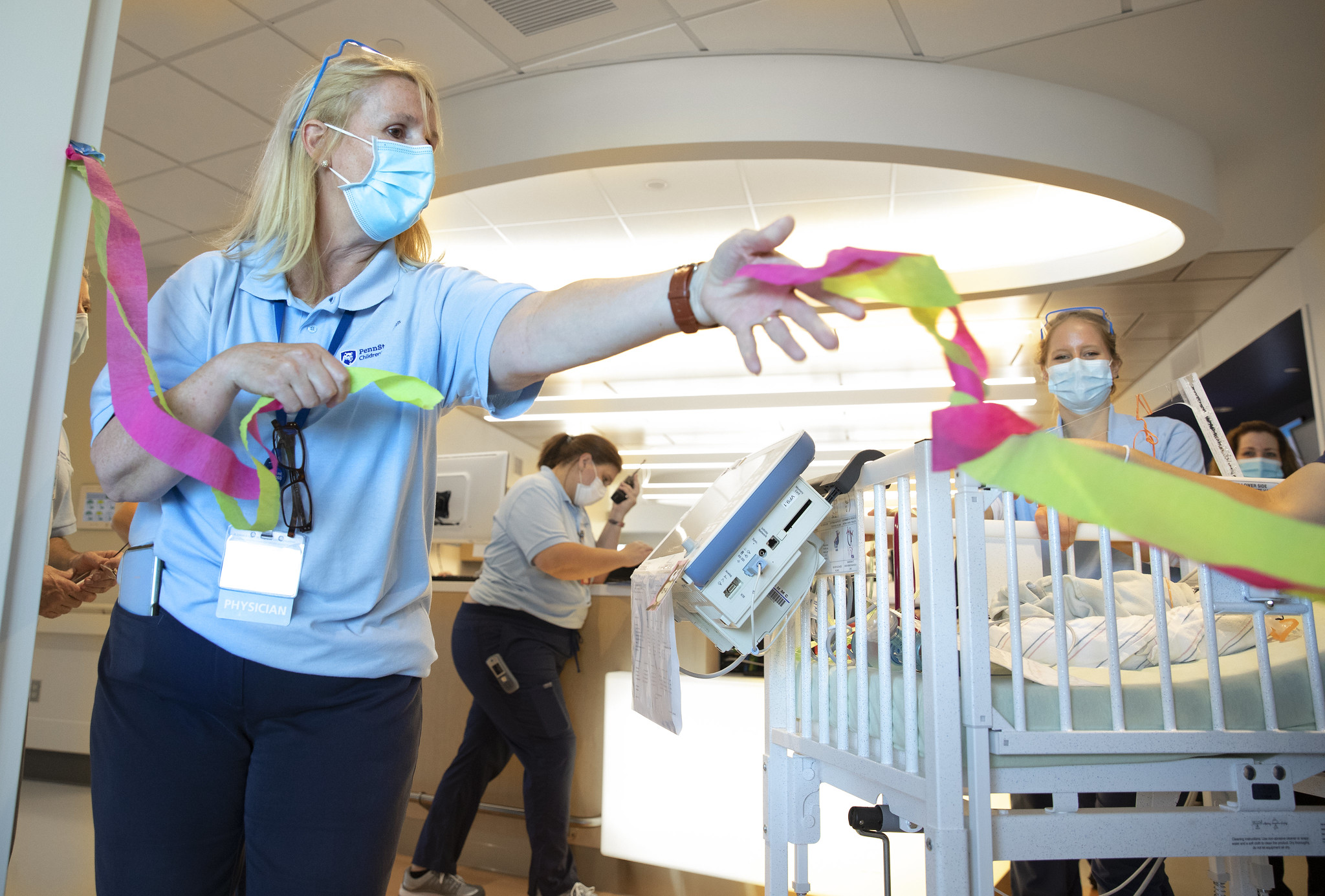 Dr. Sarah Iriana extends her arm holding a handful of ribbon next to a crib. Equipment is draped from the side of the crib. Behind her, three women in surgical masks look on or focus on other work.