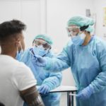 Two health care workers in personal protective equipment administer a COVID-19 test on a patient.
