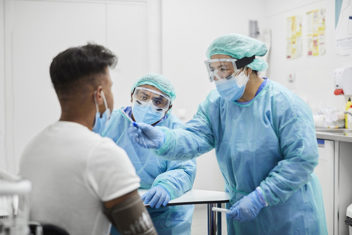 Two health care workers in personal protective equipment administer a COVID-19 test on a patient.