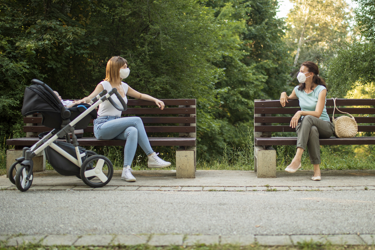 Two women wearing facemasks sit outside on separate benches, facing each other. One woman has her hand on a nearby stroller.