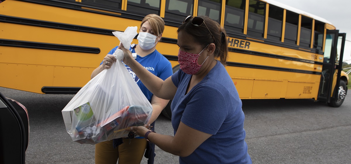 Two women hold a trash bag filled with school supplies. They are both wearing face masks. Jessica Wadsworth, on the left, is wearing a Penn State Health T-shirt. Behind them is a school bus.