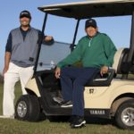 One man sits in and another stands next to a golf cart in a grassy area on a sunny day.