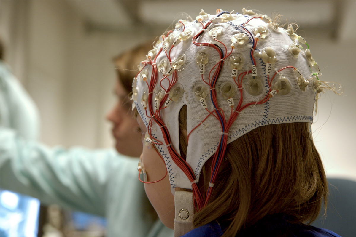 A female wears a rubber treatment cap lined with electrodes as a medical professional stands nearby.