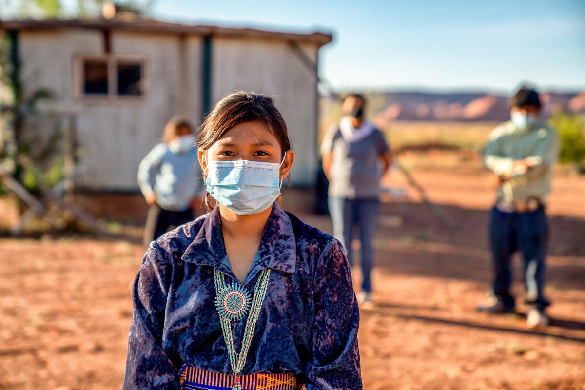 Native American teen-aged girl wears a face mask and a beaded necklace. Standing behind her are three other people wearing face masks near a building.