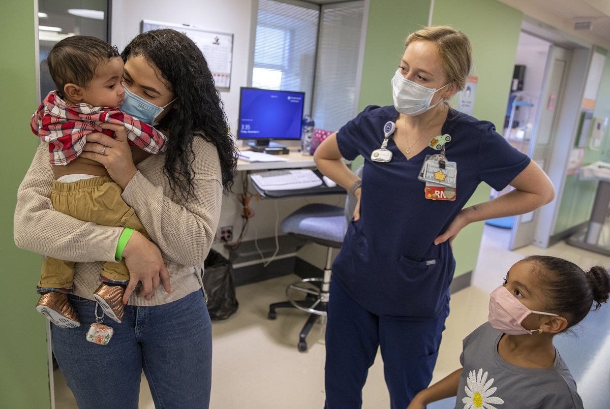 Terianny Vicente holds son Calvin, who is wearing a plaid shirt and pants, as Jo Rosenberger and Calvin’s big sister look on. Vicente, who has long hair, wears a sweater, jeans and a mask. Rosenberger wears scrubs and a mask. Hospital equipment and computer monitors are in the hallway behind them.