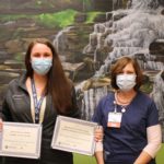 Elisha Peregino holds two plaques flanked by two of her colleagues in front of a background painting featuring waterfalls.