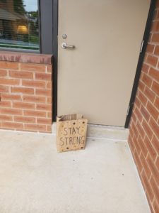 A paper bag with the words “Stay Strong” written on it sits outside a door of a brick building.