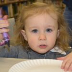 A close-up of a toddler girl shows her holding a crayon and touching a paper plate she is about to color on. She is wearing a long-sleeved top and a watch. Behind her are shelves of books and a person wearing jeans.