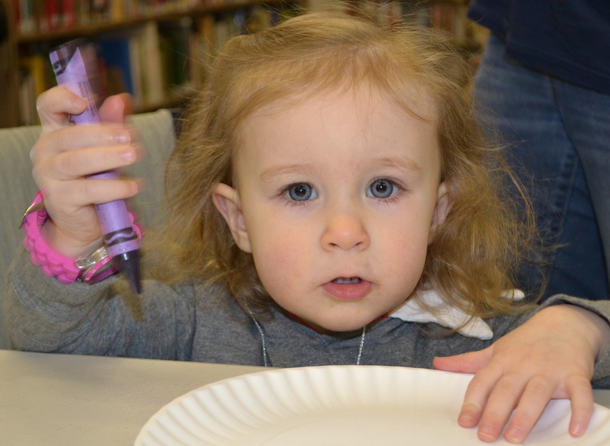 A close-up of a toddler girl shows her holding a crayon and touching a paper plate she is about to color on. She is wearing a long-sleeved top and a watch. Behind her are shelves of books and a person wearing jeans.