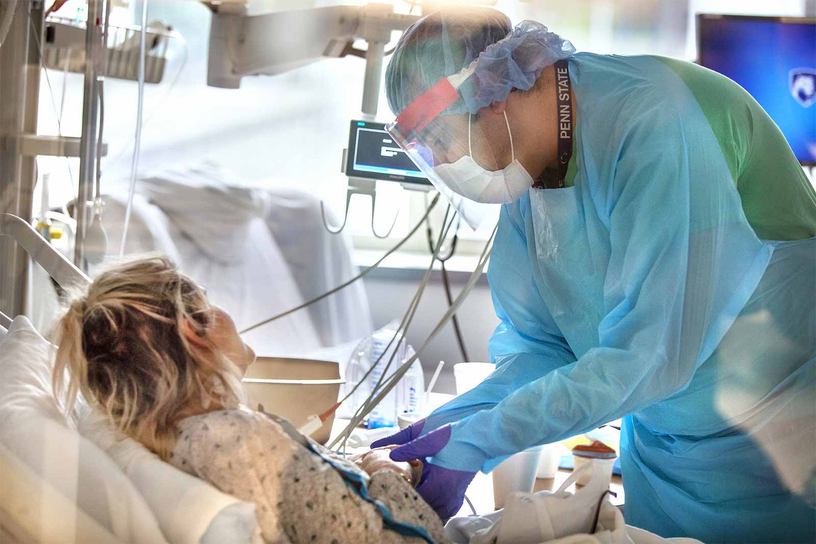 Un cuidador que lleva una bata, máscara, protector facial y guantes, sostiene la mano de una paciente que está en una cama de hospital. Un monitor y una ventana se encuentran entre los elementos en el trasfondo.
