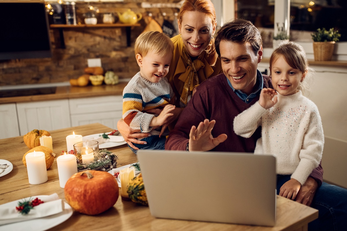 A mother, father and two young children gather around one end of a dinner table, set for the Thanksgiving holiday. A laptop is open on the table and the family is peering at the screen, waving, to show social distancing.