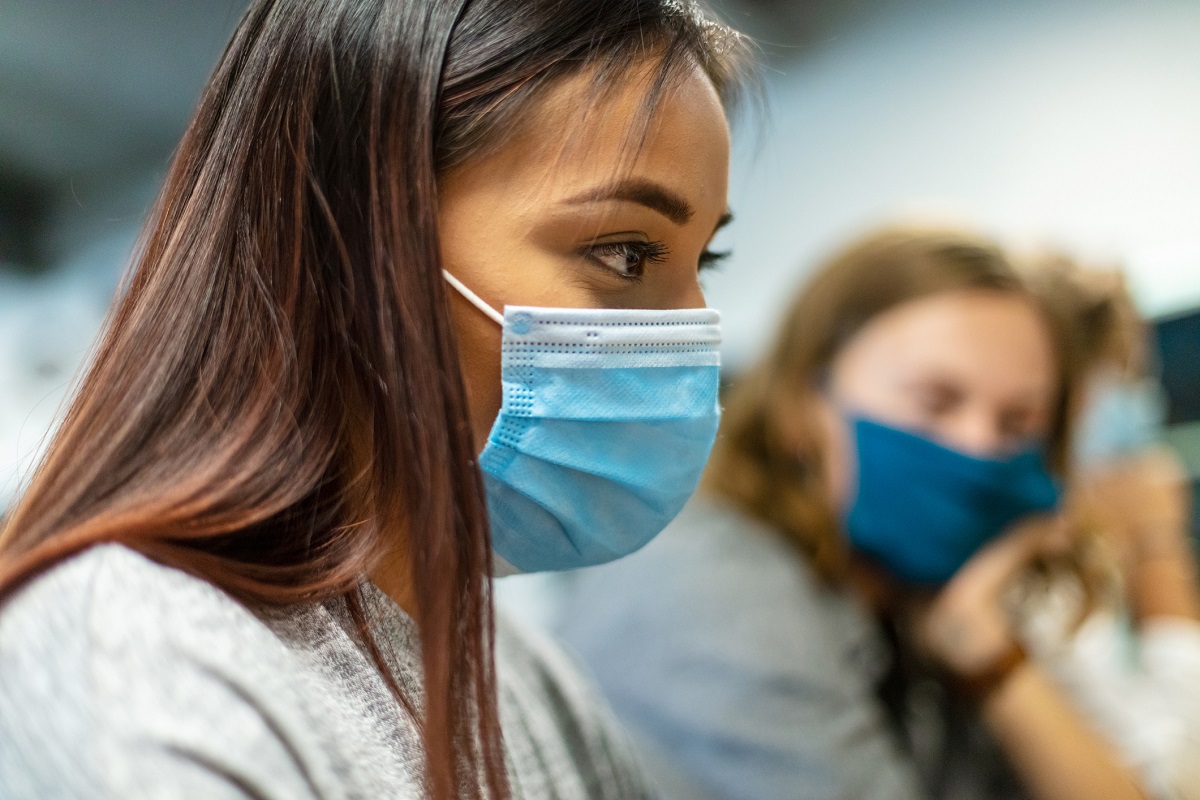 Up-close picture of a young woman looking ahead while wearing a face mask. Close by but out of focus, another woman wears a face mask and looks tired.
