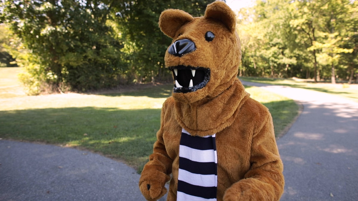Person dressed as the Penn State Nittany Lion and wearing a scarf runs along a path. Behind him are two asphalt paths, grass and trees.