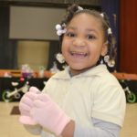 A little girl with braids in her hair smiles. She is wearing knit gloves.
