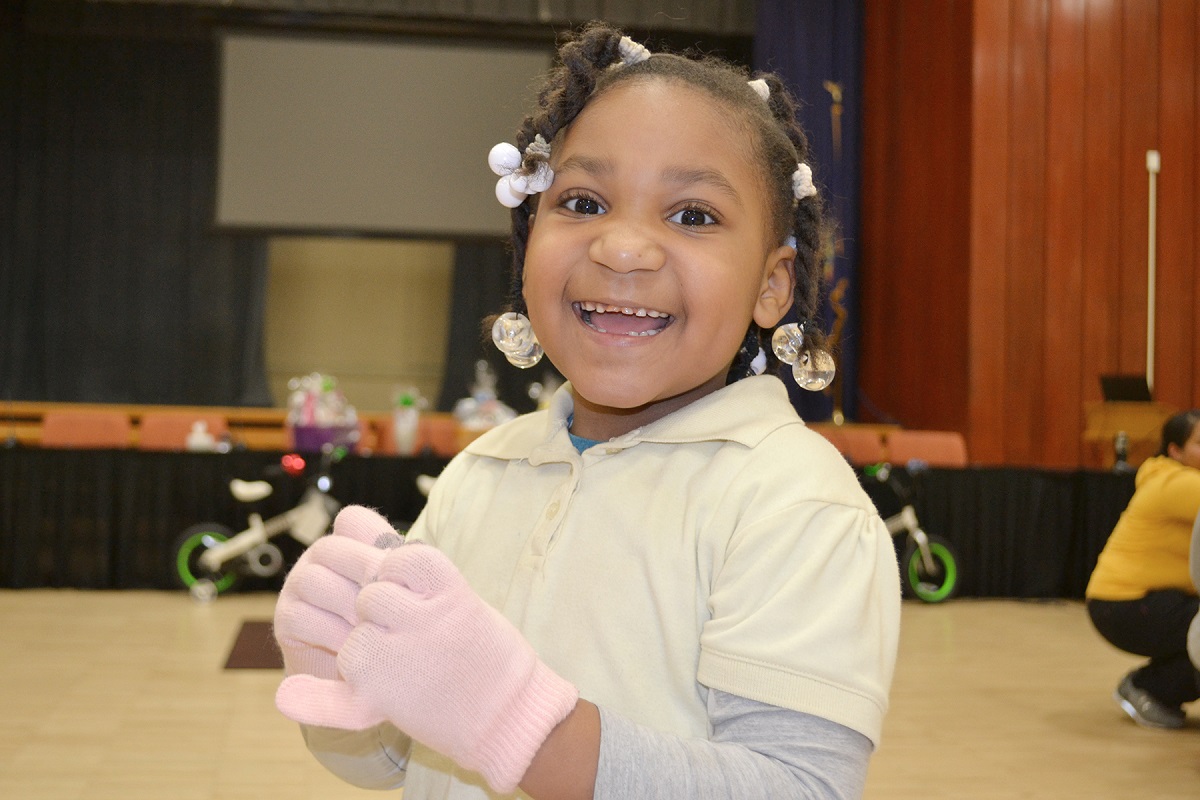 A little girl with braids in her hair smiles. She is wearing knit gloves.