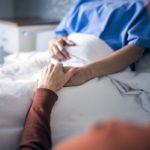 Female patient in a hospital bed, only hands and upper body visible, holding hands with a man, only hands and arms visible.
