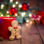 A gingerbread man leans against a mug containing a hot liquid. Behind them holiday decorations are seen out of focus.