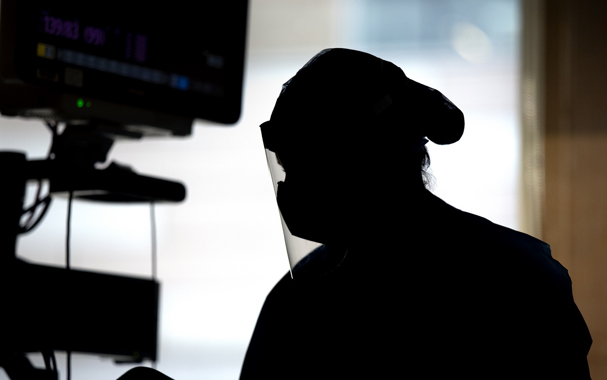 A silhouette of a medical care provider wearing protective gear, including a face shield and mask, while working in a patient room.