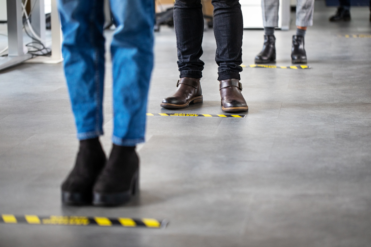 Pictured from the knees down, people stand at a series of taped-off lines on a floor.