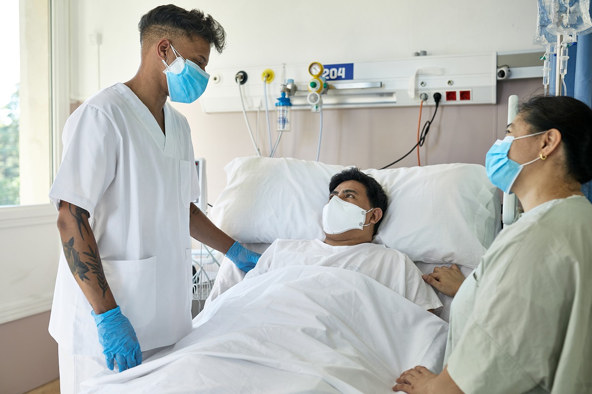 Two people stand on either side of a patient’s bed. All three are wearing face masks.