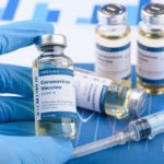 A hand wearing gloves holds a vial of coronavirus vaccine. Three vials are next to the hand. A syringe is in front of them.