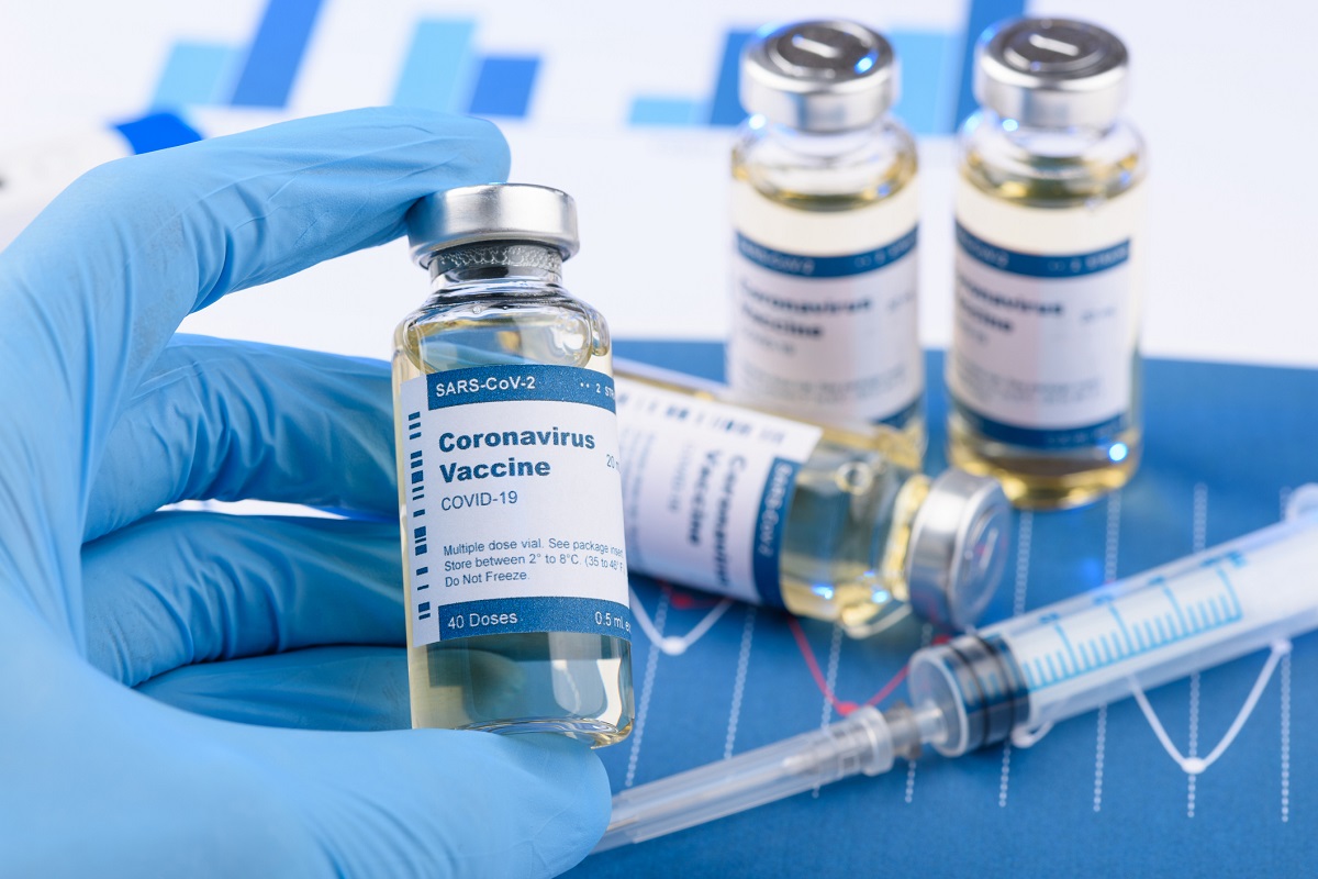 A hand wearing gloves holds a vial of coronavirus vaccine. Three vials are next to the hand. A syringe is in front of them.