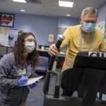 Woman in grey Penn State Health jacket wearing safety glasses, facemask and gloves, writes on a clipboard as a man in a yellow shirt wearing a face mask walks on an exercise machine during an in-person cardiac rehabilitation visit.