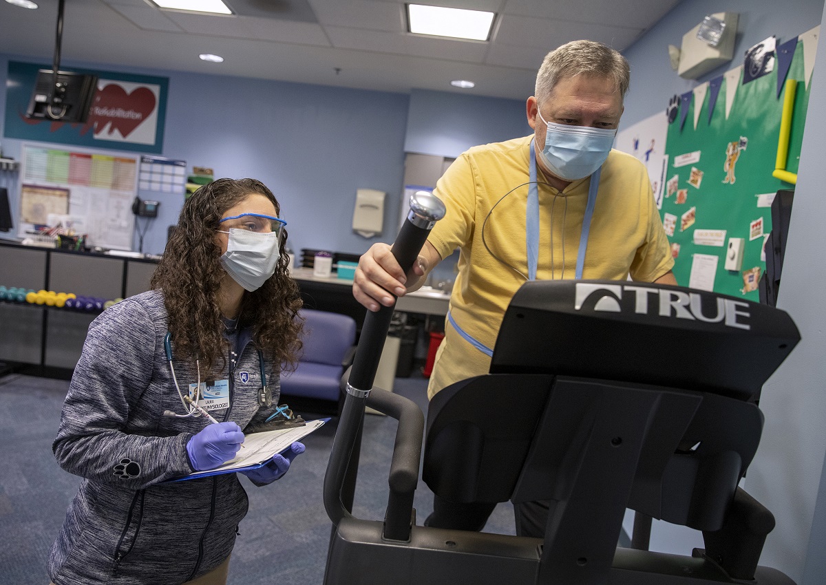 Woman in grey Penn State Health jacket wearing safety glasses, facemask and gloves, writes on a clipboard as a man in a yellow shirt wearing a face mask walks on an exercise machine during an in-person cardiac rehabilitation visit.