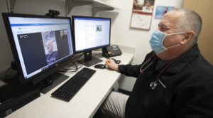 Man seated wearing a black jacket and a facemask with a stethoscope around his neck smiles at his computer screen. Man with blue shirt and grey beard is smiling on screen during a virtual hybrid cardiac rehabilitation appointment.