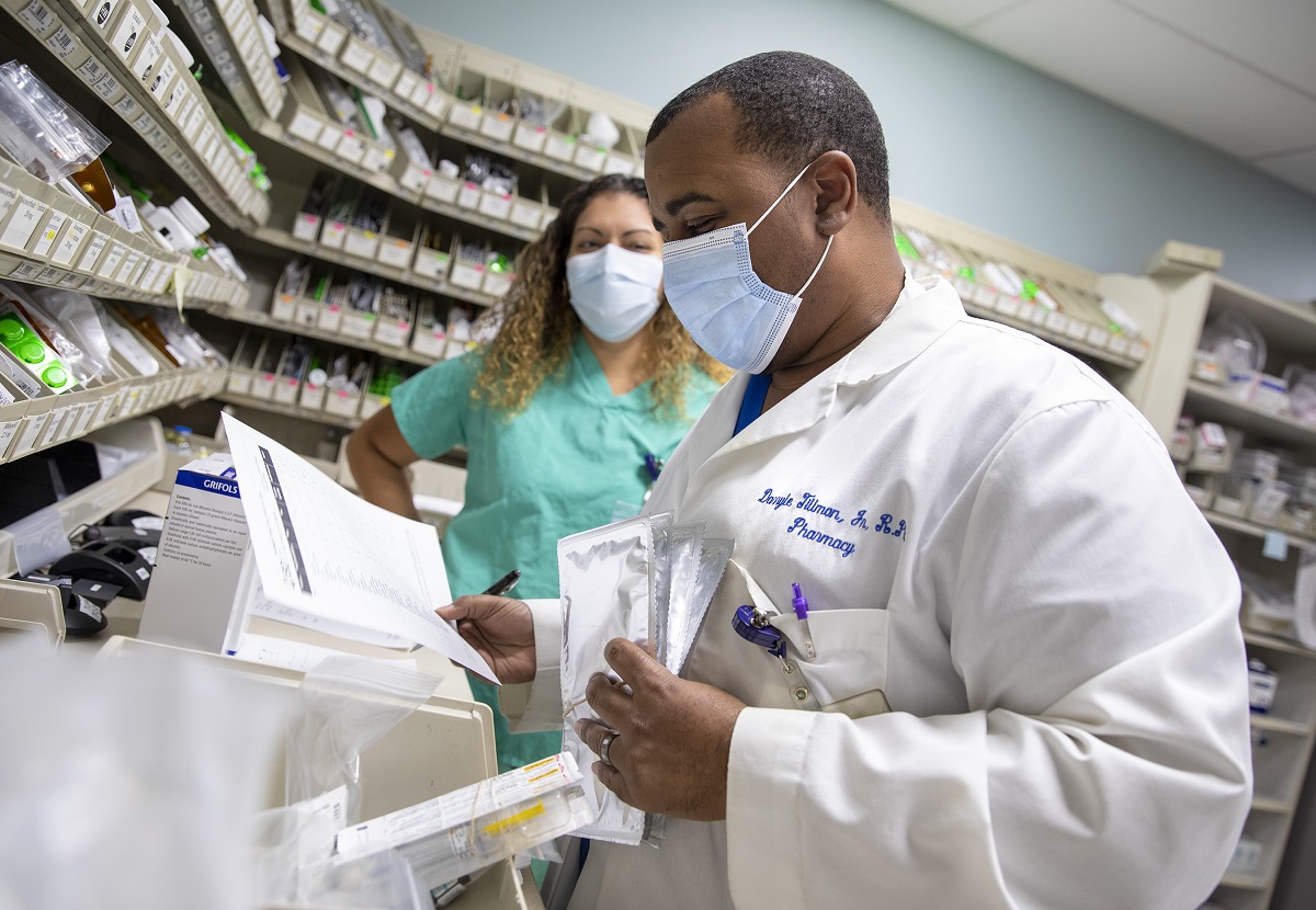 Darryle Tillman, a pharmacist at Penn State Health St. Joseph Medical Center, holds several papers and foil packets with medicine. He is wearing a face mask and white coat with his name and the word “Pharmacy” on it. Behind him is technician Karla Rivera-Rodriguez who is wearing scrubs and a face mask and has long, curly hair.