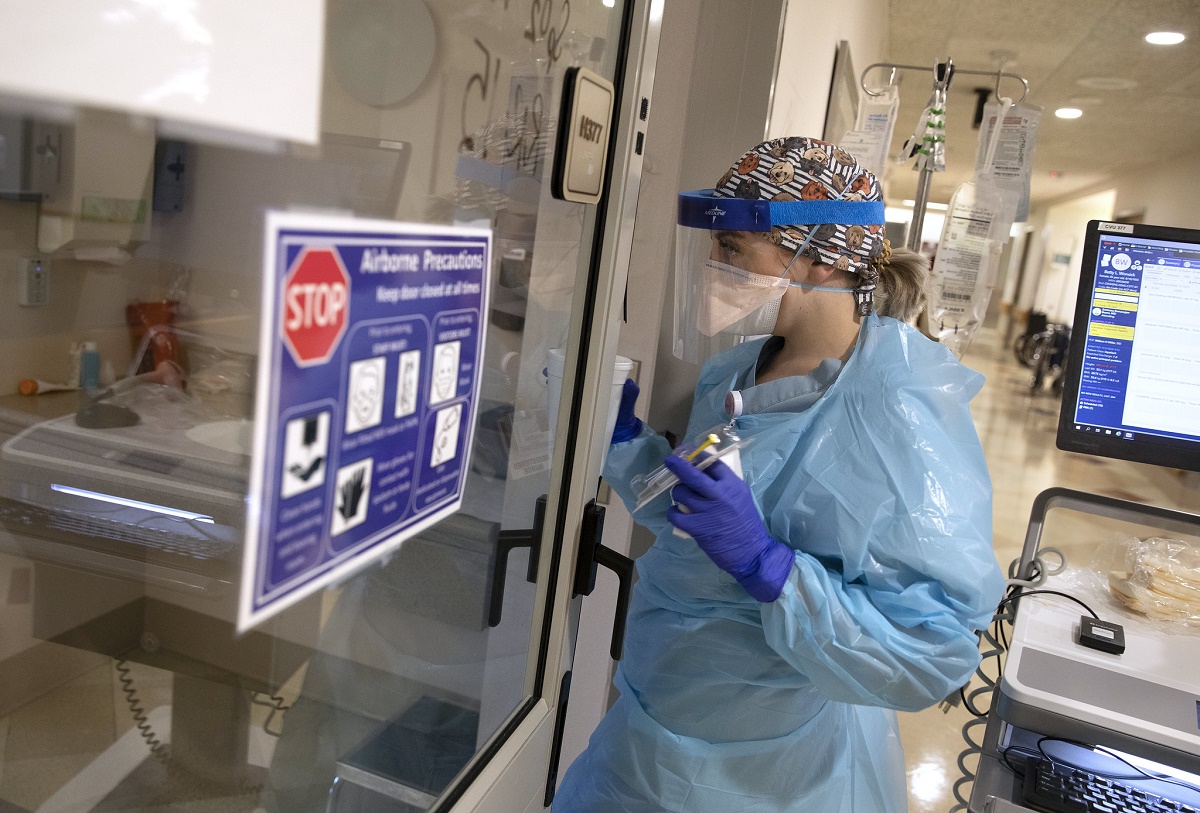 Nurse wearing protective blue gown, face mask and eye shield holds medication and looks into window of COVID-19 patient’s room.