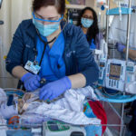 A medical worker provides care to a baby in the NICU while another worker looks on in the near background. An IV pole is also in the photo.