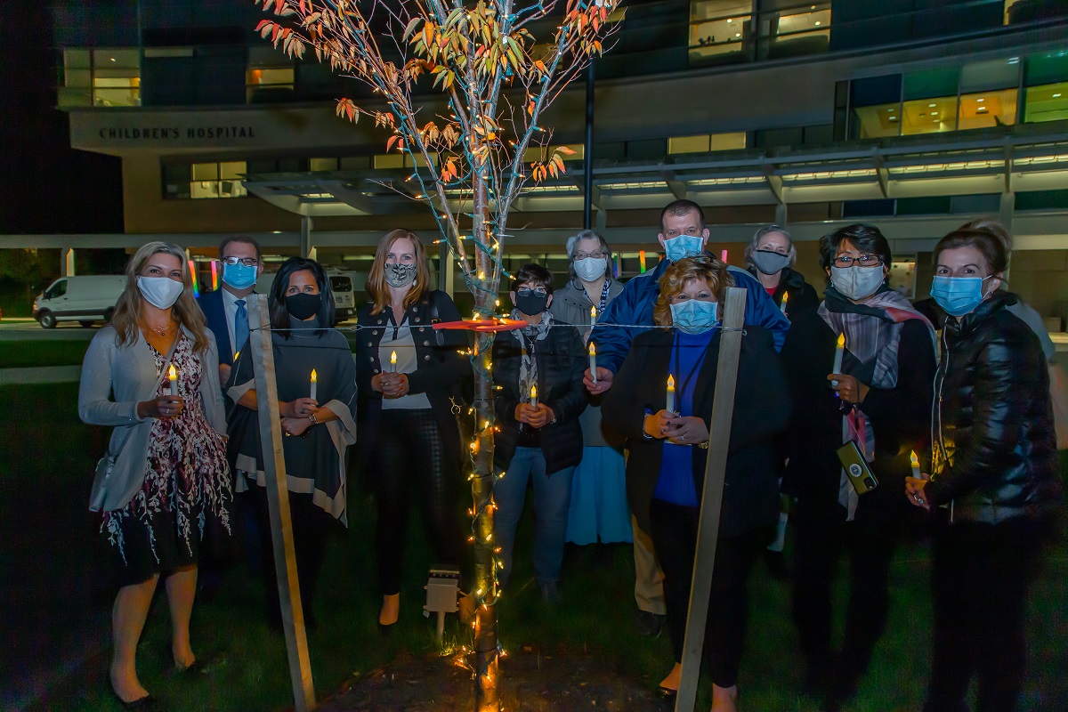 A group of 12 people stand behind a small tree decorated in lights at night. They are holding lit, battery-operated candles. Behind them is Penn State Health Children's Hospital.