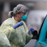 A nurse in full personal protective equipment administers a COVID-19 test at a drive-through testing location