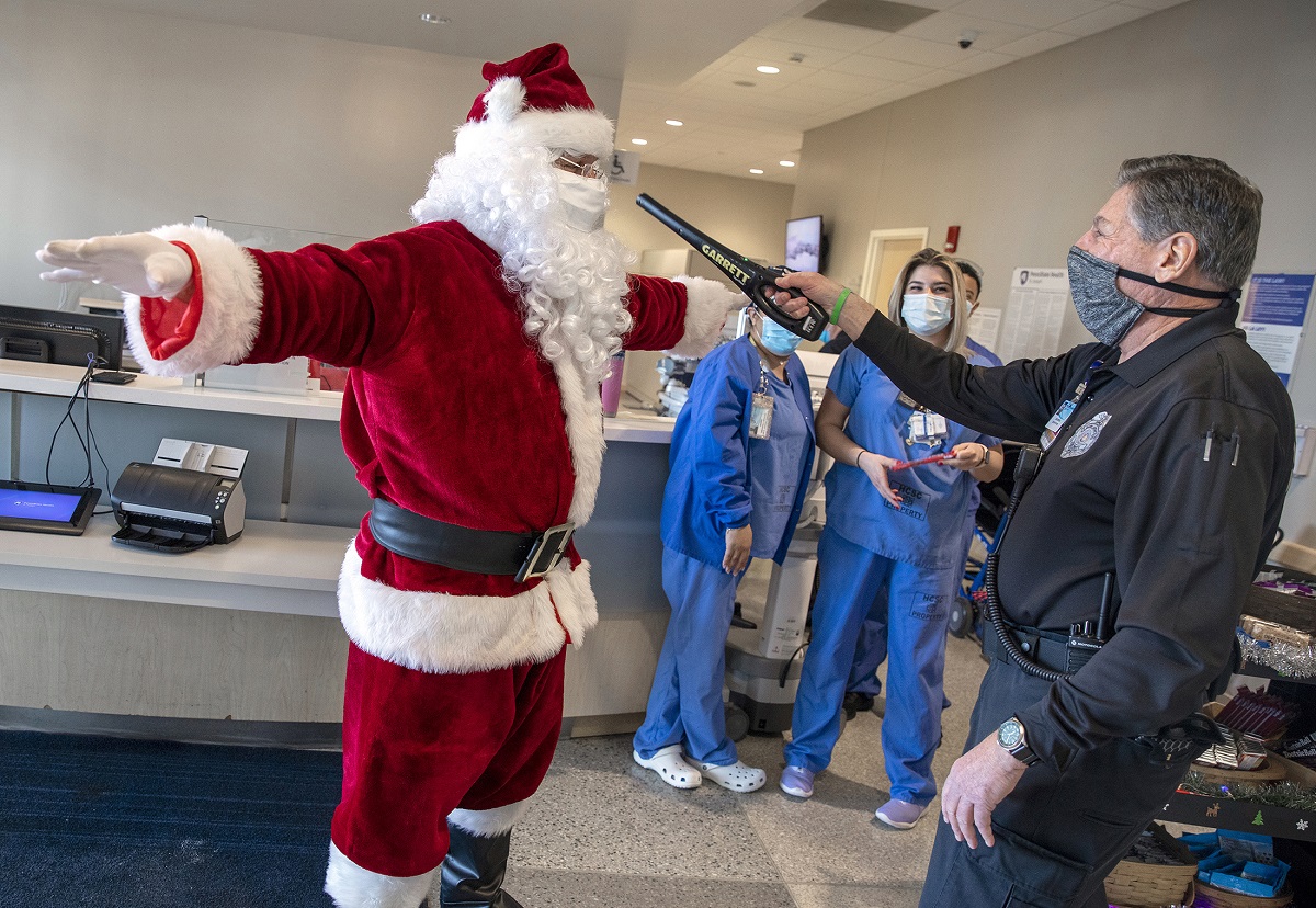 A man in a Santa Claus suit is wanded by a security guard while people in scrubs and surgical masks look on.