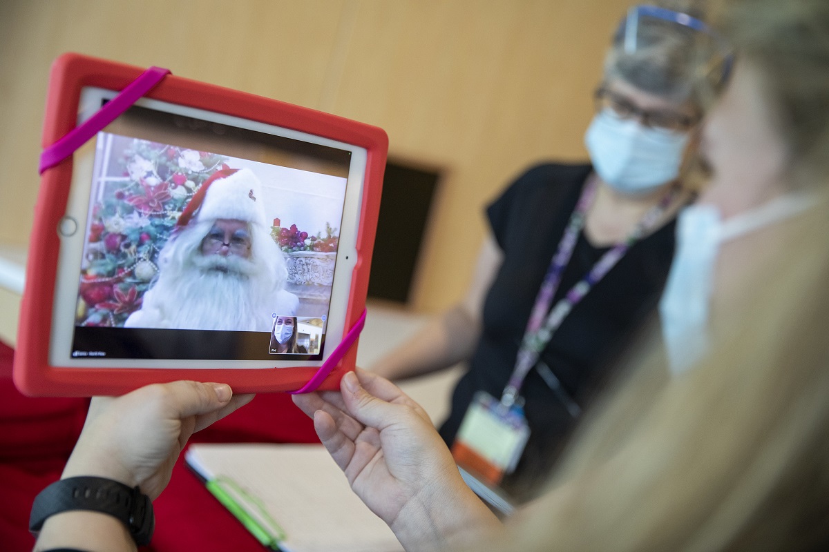 A woman holds a notepad, upon which is a view of Santa Claus’s face. Next to her is another woman in personal protective equipment.