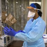 Female doctor in scrubs, face shield and N95 mask adjusts her blue surgical gloves. Behind her, brown paper bags labeled with last names and dates hang from the wall. Used face masks are stored in the bags.