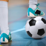 Close-up of an athlete’s legs as they stand on a gym floor, the right foot resting on a soccer ball.