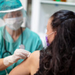 A female medical staff member wearing scrubs, a mask, a face shield and a stethoscope wipes the left arm of a female patient, who faces away from the camera.
