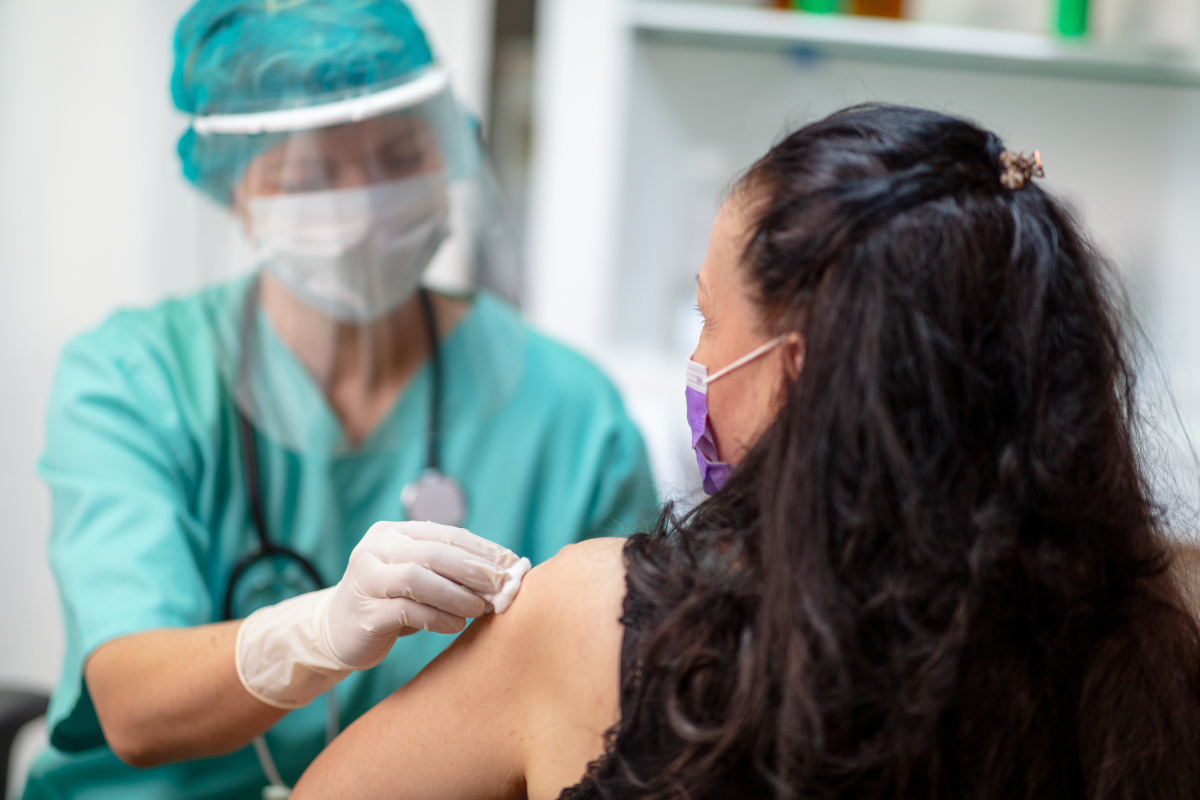 A female medical staff member wearing scrubs, a mask, a face shield and a stethoscope wipes the left arm of a female patient, who faces away from the camera.