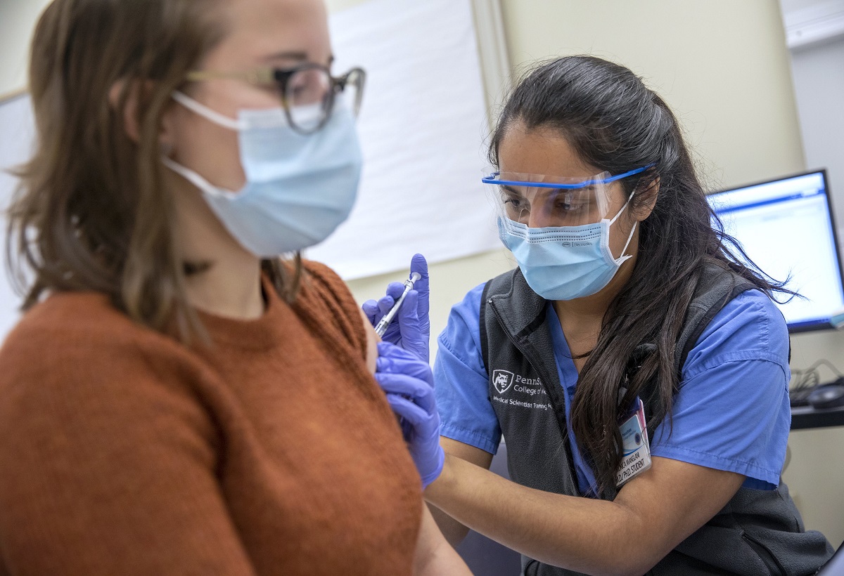 A young graduate student wearing eye protection, a surgical mask and blue gloves administers a shot into the upper arm of a young woman, wearing a mask, in the foreground.