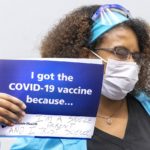 A female health care worker, wearing a face mask and eye protection, winks at the camera while holding a sign that reads, “I got the COVID-19 vaccine because I’m a single parent and I trust science.”