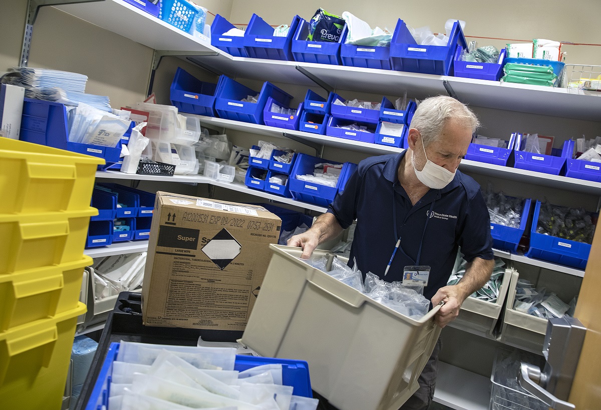 Stephen Caldwell, an inventory management specialist at Penn State Health Lime Spring Outpatient Center, lifts a box with medical supplies. He is wearing a polo shirt with the Penn State Health logo on it, a name badge and a face mask. Behind him are four shelves with supplies. Large plastic boxes are stacked to the left of him.