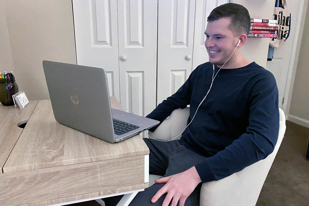 A man sits at a desk, wearing earbuds, looking at a laptop screen. He smiles slightly. A door, bookshelf and closet are in the background.